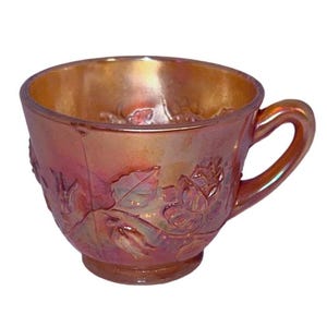 May include: Iridescent orange teacup with a handle and a floral relief pattern. The cup has a round base and a wide opening. The floral design includes roses and leaves. The cup is made of glass.