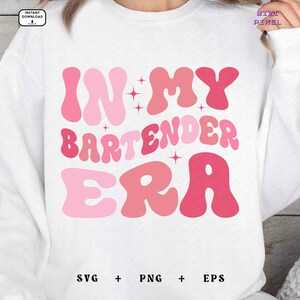 May include: White sweatshirt with pink text that reads "In My Bartender Era" in a retro font. The text is surrounded by pink stars.