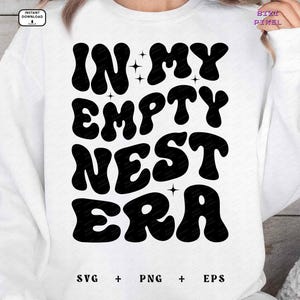 Op de afbeelding: Wit sweatshirt met zwarte tekst die "In My Empty Nest Era" luidt in een retro lettertype.