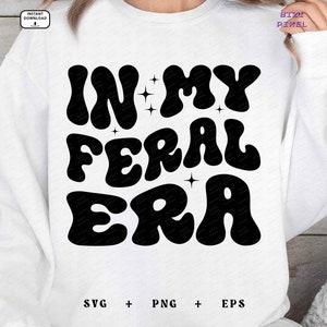 May include: White sweatshirt with black text that reads "In My Feral Era" in a retro font. The text is surrounded by small stars.