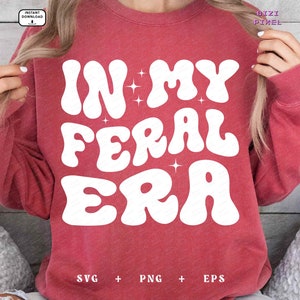 May include: Red sweatshirt with white text that reads "In My Feral Era" in a retro font. The text is surrounded by small white stars.