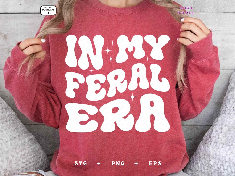 May include: Red sweatshirt with white text that reads "In My Feral Era" in a retro font. The text is surrounded by small white stars.