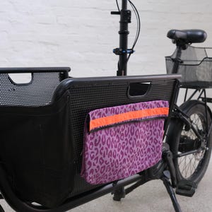 Könnte beinhalten: Schwarzes Lastenrad mit einer lila Tasche mit Leopardenmuster. Die Tasche hat einen orangefarbenen Streifen oben. Das Fahrrad hat einen schwarzen Rahmen, Lenker und einen Korb.