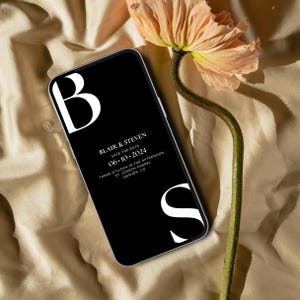 May include: A black smartphone with a white 'B' and 'S' on the screen. The screen displays a wedding save the date announcement for Blair & Steven on June 10, 2024, at 3:00 PM at St. Joseph Chapel in Denver, CO.