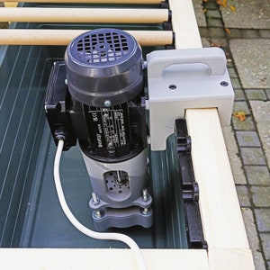 Könnte beinhalten: Ein grauer Elektromotor mit schwarzem Lüfterabdeckung und weißem Stromkabel. Der Motor ist an einem Holzrahmen mit einer grauen Kunststoffhalterung befestigt. Die Halterung hat einen Griff.