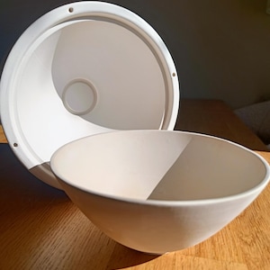 Puede incluir: Un bol de cerámica blanca con un interior liso y redondeado. El bol está sobre una superficie de madera.