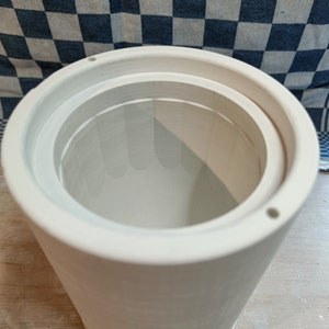 Close-up of a cylindrical plaster mold with internal steps and small holes around the top edge. A blue and white checkered cloth is visible in the background.