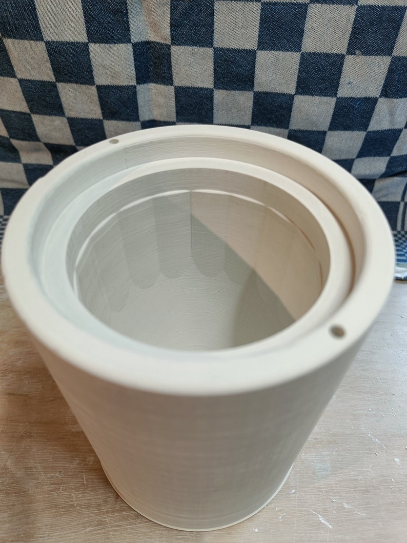 Close-up of a cylindrical plaster mold with internal steps and small holes around the top edge. A blue and white checkered cloth is visible in the background.