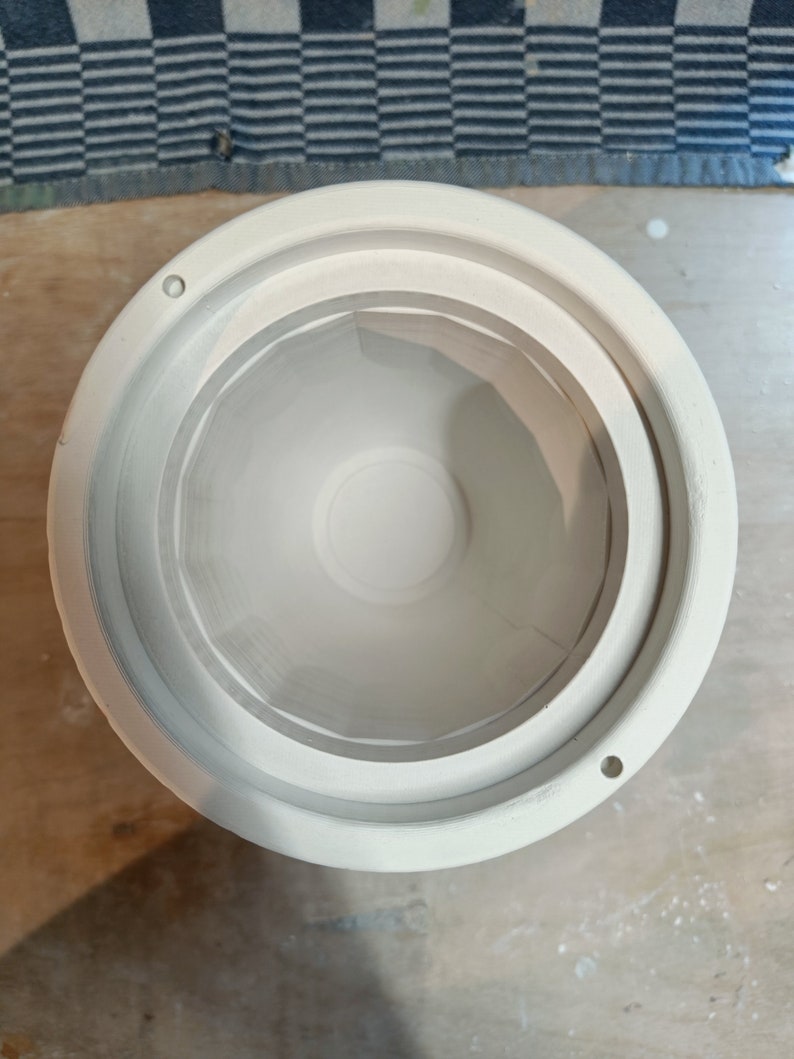 Top-down view of a white plaster mold cylinder with interior steps and small holes. Blue and white checkered fabric in the background.