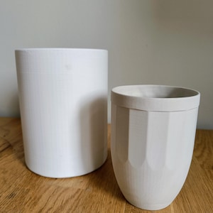 Pode incluir: Dois vasos de cerâmica branca, um alto e cilíndrico, o outro mais baixo e com um design canelado. Ambos os vasos estão sobre uma superfície de madeira.
