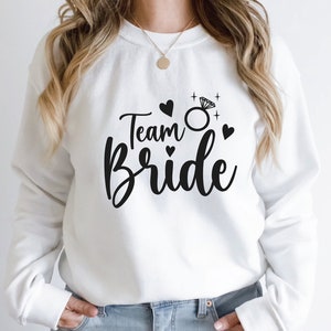 Könnte beinhalten: Weißer Pullover mit dem Text "Team Bride" in schwarzer Schrift. Ein Diamantring mit einem Glitzereffekt befindet sich über dem Wort "Bride". Um den Ring und den Text sind kleine Herzen angeordnet.