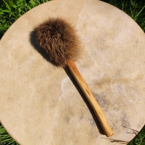 Op de afbeelding: Een ronde, lichtbruine trommel met een bruine, met bont bedekte klopper. De trommel ligt op groen gras en de klopper heeft een lichtbruine houten steel. Het oppervlak van de trommel heeft een natuurlijk, getextureerd uiterlijk.
