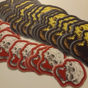 May include: A stack of black and yellow skull stickers with a white outline. The stickers are layered on top of each other, creating a 3D effect. Below the stack are several red and white skull stickers with a black outline.