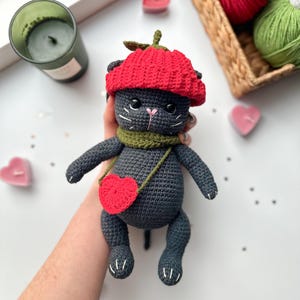 May include: A handmade crochet cat doll in dark gray, wearing a red beret and a green scarf. The cat has a red heart-shaped bag and white details on its paws. The doll is held in a hand, with a candle and yarn in the background.