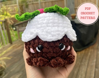 Könnte beinhalten: Gehäkelter Weihnachtspudding mit weißem Oberteil und brauner Basis. Er hat grüne Stechpalmenblätter und ein niedliches Gesicht mit schwarzen Augen und rosa Wangen. Der Text "PDF Crochet Pattern" ist enthalten.