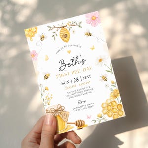 May include: A rectangular birthday invitation card featuring watercolor illustrations of bees, honeycombs, flowers, and a honey jar. The text reads: JOIN US TO CELEBRATE Beth's FIRST BEE-DAY SUN 28 MAY 2:00 PM 4:00 PM SMITH'S RESIDENCE 1234 BIRTHDAY AVENUE ORLANDO, FLORIDA RSVP CHARLOTTE SMITH AT 123-456-7890.