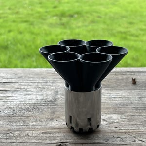 Pode incluir: Um objeto de metal preto e prateado com sete aberturas em forma de cone. O objeto tem uma base cilíndrica com detalhes recortados. Está sobre uma superfície de madeira desgastada com um fundo de relva verde.