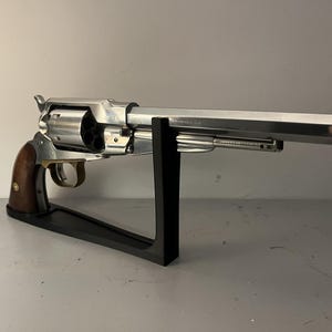Könnte beinhalten: Ein silberfarbener Revolver mit Holzgriff wird auf einem schwarzen Ständer präsentiert. Die Schusswaffe hat einen langen Lauf und eine Trommel mit mehreren Kammern. Der Griff ist dunkelbraun und das Metall hat eine polierte Oberfläche.