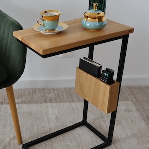 Könnte beinhalten: Ein schwarzer Metall-Beistelltisch mit einer Holzplatte und einem Ablagefach. Der Tisch hat ein geschwungenes Design und steht neben einem dunkelgrünen Sessel.