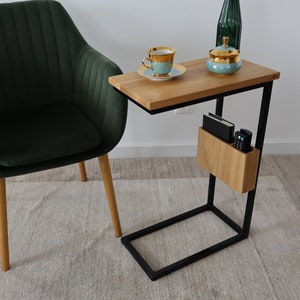 May include: A dark green velvet armchair with wooden legs sits next to a black metal side table with a wooden top. The table has a small shelf for storage and a cup of tea and a sugar bowl are on the top.