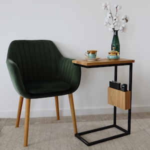 May include: A dark green velvet armchair with wooden legs sits next to a black metal side table with a wooden top. The table has a shelf for storage and a small wooden box on the shelf. A white cotton flower arrangement is in a green glass vase on the table.