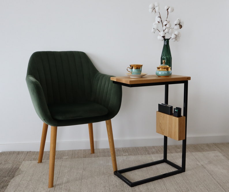May include: A dark green velvet armchair with wooden legs sits next to a black metal side table with a wooden top. The table has a shelf for storage and a small wooden box on the shelf. A white cotton flower arrangement is in a green glass vase on the table.