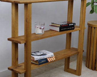 Peut inclure: Une console en bois brun clair dotée de trois étagères horizontales. L'étagère supérieure supporte une lampe blanche, un vase blanc avec des fleurs violettes et un cadre photo. L'étagère centrale contient une bougie en verre et deux livres.