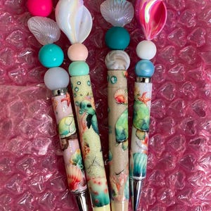 Puede incluir: Cuatro bolígrafos decorativos con diseños marinos. Cada bolígrafo presenta una combinación única de cuentas coloridas, conchas y criaturas marinas. Los barriles de los bolígrafos están adornados con ilustraciones de tortugas, conchas y corales, con detalles plateados y amarillos.