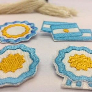 Op de afbeelding: Vier geborduurde patches met de zon en een blauw-wit ontwerp. Twee patches zijn rond met een gele zon in het midden. Eén patch is een rechthoekige vlag met een blauw-wit ontwerp en een gele zon in het midden. De vierde patch is een rond ontwerp met een gele zon in het midden en een blauw-witte rand.