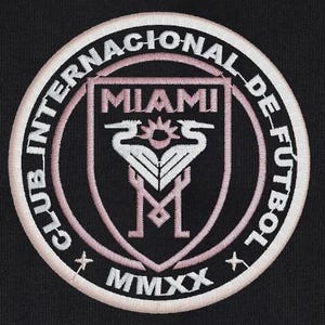 May include: A pink and white embroidered patch with the text "Club Internacional de Futbol Miami MMXX". The patch features a crest with two white birds and the word "Miami" in pink.