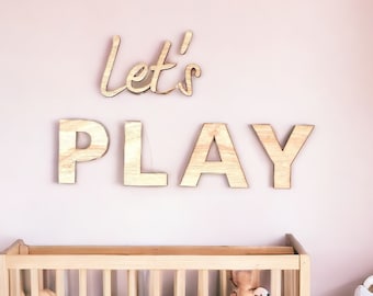 May include: Wooden wall decor with the words "Let's Play" in a nursery setting with a crib and a bassinet.