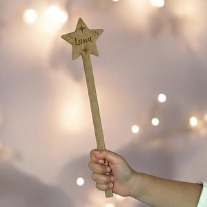 May include: A wooden star-shaped wand with the name "Luna" engraved on it. The wand is held by a child's hand against a blurred background of white lights.
