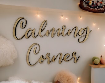 Può includere: Decorazione da parete in legno con la scritta "Calming Corner" in corsivo. Le lettere sono marrone chiaro con un contorno nero, montate su una parete di colore chiaro. Luci decorative e una piccola mensola con peluche sono visibili.
