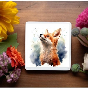 May include: A square white coaster featuring a watercolor painting of a fox looking upwards. The fox has orange and white fur with blue and green watercolor accents. The coaster is surrounded by colorful flowers.