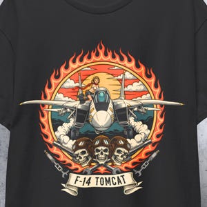 May include: Black t-shirt featuring a graphic of an F-14 Tomcat fighter jet with a pilot, surrounded by flames and skulls. The text "F-14 TOMCAT" is displayed on a banner below the image. The design is in shades of orange, white, and blue.