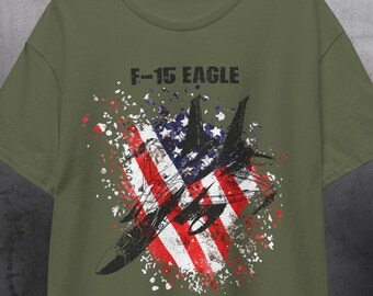 Puede incluir: Camiseta verde oliva con un gráfico de un avión de combate F-15 Eagle y un diseño de bandera estadounidense. El diseño incorpora rayas rojas y blancas, un campo de estrellas azul y el texto "F-15 EAGLE" encima del avión.