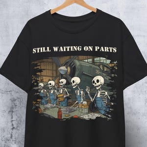 以下が含まれることがあります： 黒いTシャツ。オーバーオールを着た骸骨が飛行機を修理しているユーモラスなイラスト。画像の上には「STILL WAITING ON PARTS」の文字がプリントされています。背景には格納庫とヴィンテージの飛行機が描かれています。