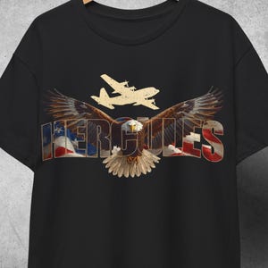 Pode incluir: T-shirt preto com um gráfico de uma águia com as asas abertas, a palavra "HERCULES" em cores patrióticas e uma silhueta de avião. O design incorpora elementos da bandeira americana.