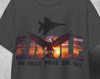 May include: Dark gray t-shirt featuring a majestic eagle with outstretched wings against a sunset backdrop. The word "EAGLE" is prominently displayed, along with the phrase "THE EAGLE RULES THE SKY" and a silhouette of a jet.