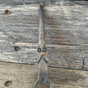 May include: A rustic, hand-forged metal bottle opener. The opener features a stylized face design with a star detail and a loop at the top. The metal has a weathered, antique appearance.