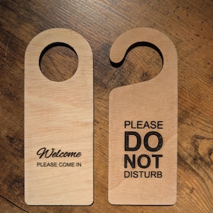 May include: Two wooden door hangers. One has a circular opening and the text "Welcome PLEASE COME IN" in black. The other has a curved top and the text "PLEASE DO NOT DISTURB" in black. Both are made of light-coloured wood.