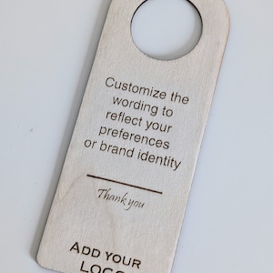 Op de afbeelding: Een houten deurhanger met een gat bovenaan om op te hangen. De hanger is leeg en heeft de tekst "Pas de tekst aan om uw voorkeuren of de identiteit van uw merk weer te geven" en "Bedankt" erop gedrukt. De onderkant van de hanger heeft de tekst "VOEG UW LOGO TOE" erop gedrukt.