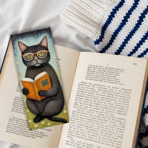 May include: A black cat wearing round glasses sits on a bed of green grass and reads a book. The cat is wearing a black and white striped collar. The background is a blue sky with stars.