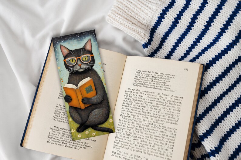 May include: A black cat wearing round glasses sits on a bed of green grass and reads a book. The cat is wearing a black and white striped collar. The background is a blue sky with stars.