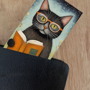 May include: A black cat wearing orange glasses is reading a book. The cat is sitting in front of a blue background with white stars. The book is orange and has a gold design on the cover.