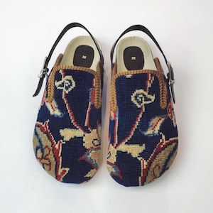 Könnte beinhalten: Ein Paar Slipper mit dunkelblauem Textilobermaterial mit mehrfarbigem Blumenmuster, beigefarbener geflochtener Verzierung und einem schwarzen, verstellbaren Fersenriemen mit silberner Schnalle. Das helle Fußbett ist mit der Größe 38 gekennzeichnet.