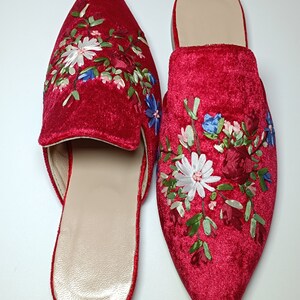 May include: A pair of red velvet slip-on shoes with pointed toes. The shoes are embroidered with a floral design in white, blue, and green. The interior of the shoes is a light beige color. The shoes are displayed on a white surface.