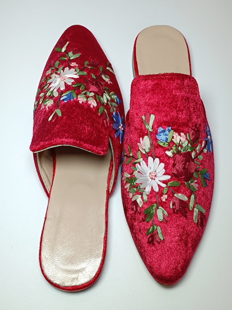 May include: A pair of red velvet slip-on shoes with pointed toes. The shoes are embroidered with a floral design in white, blue, and green. The interior of the shoes is a light beige color. The shoes are displayed on a white surface.