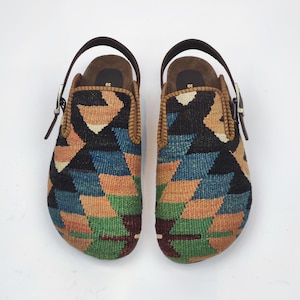 Könnte beinhalten: Ein Paar Clogs mit einem geometrischen Muster in Braun-, Blau-, Grün- und Schwarztönen. Die Clogs haben einen braunen Lederriemen und eine Schnalle sowie eine braune Lederinnensohle. Größe 39.