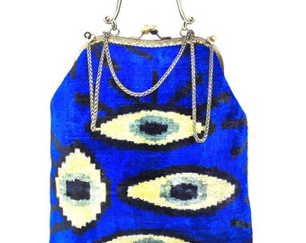 May include: A vibrant blue handbag featuring a repeating eye pattern in black and cream. The bag has a metallic gold-tone frame with a decorative floral clasp and a thin gold-tone chain handle. The fabric has a soft, textured appearance.